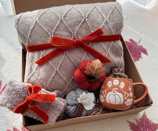 An autumn-themed gift box filled with cozy fall items, including a large beige diamond-knit blanket tied with a red ribbon, a matching knit scarf, a terracotta mug with a white pumpkin design, and small decorative pumpkins and leaves.